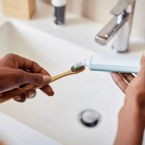 Eine Hand hält eine Zahnbürste mit grünen Borsten, während eine andere Hand eine Tube Zahnpasta hält. Die Zahnpasta wird auf die Zahnbürste gedrückt. Im Hintergrund ist ein Waschbecken und eine Wasserhahn sichtbar.