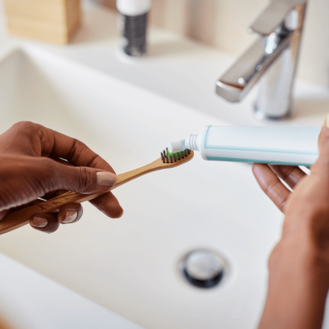 Eine Hand hält eine Zahnbürste mit grünen Borsten, während eine andere Hand eine Tube Zahnpasta hält. Die Zahnpasta wird auf die Zahnbürste gedrückt. Im Hintergrund ist ein Waschbecken und eine Wasserhahn sichtbar.
