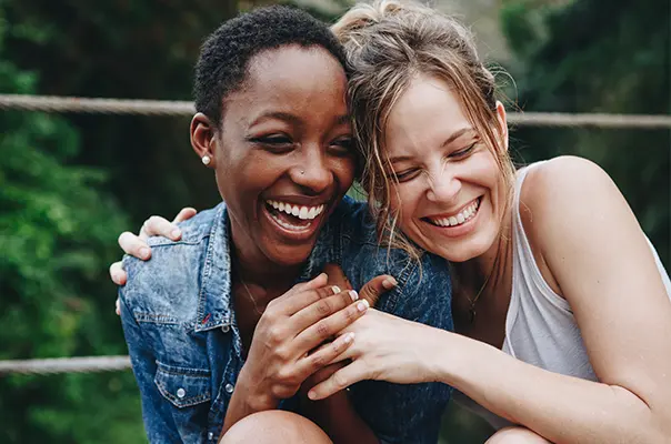 Zwei Frauen lachen fröhlich zusammen, während sie eng nebeneinander sitzen. Sie tragen lässige Kleidung und scheinen einen glücklichen Moment zu genießen. Im Hintergrund sind unscharfe grüne Bäume zu sehen, was eine natürliche Umgebung andeutet.