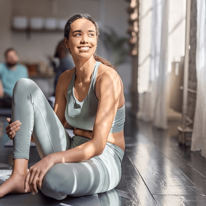Eine lächelnde Frau sitzt in Sportkleidung auf dem Boden, umgeben von indirektem Licht. Sie hat lässige, entspannte Körperhaltung und schaut optimistisch in die Ferne. Der Raum wirkt freundlich und einladend.
