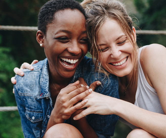 Zwei Frauen lachen herzlich und umarmen sich. Die eine trägt eine Jeansjacke, die andere ein helles Top. Ihre Gesichtsausdrücke zeigen Freude und Verbundenheit, vor einem unscharfen, grünen Hintergrund.
