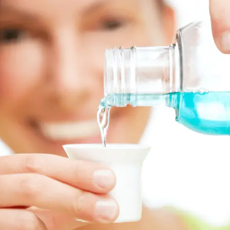 Eine Person gießt blaues Mundwasser aus einer Flasche in einen kleinen, weißen Becher. Die Person lächelt, während sie den Becher hält, bereit, das Mundwasser zu verwenden.
