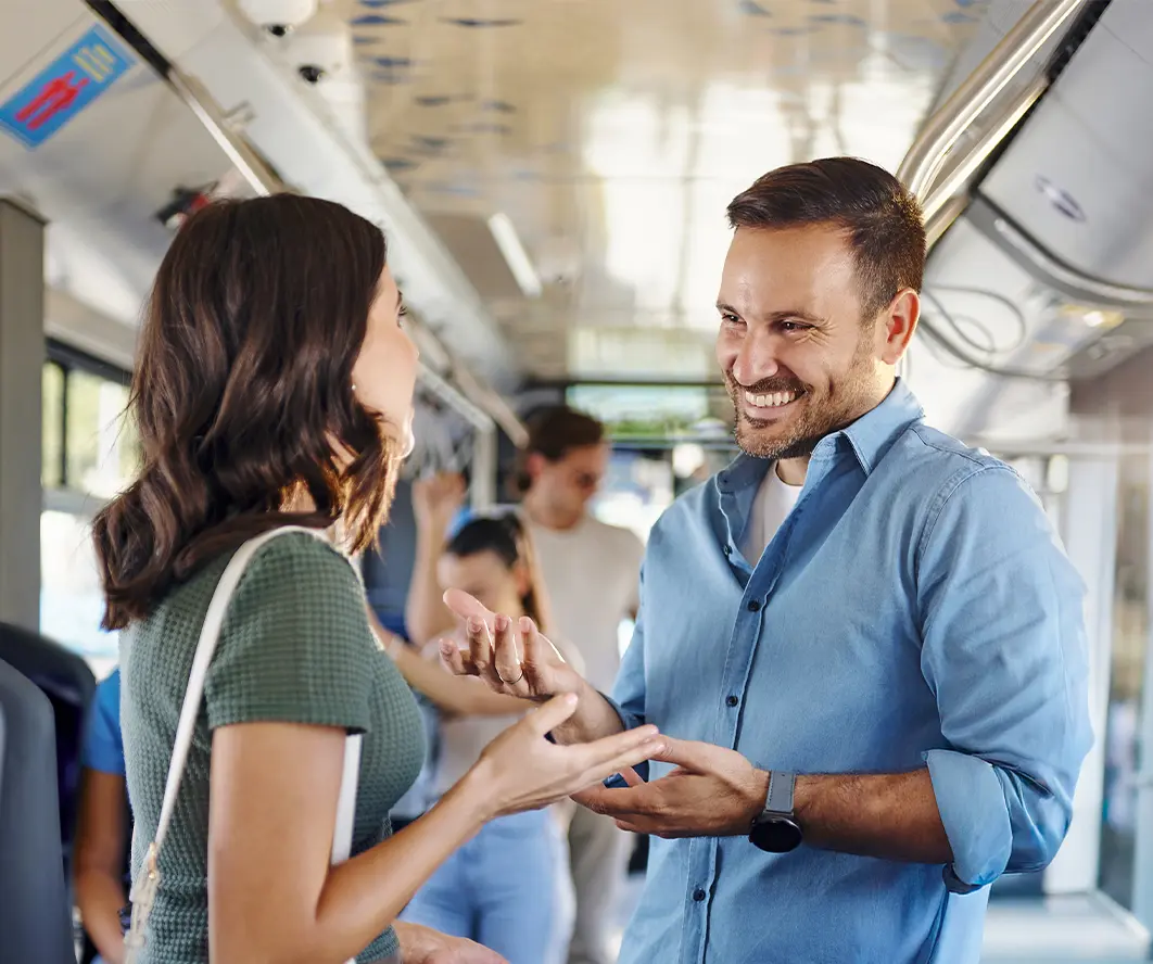 Zwei Personen unterhalten sich in einem Bus. Der Mann trägt ein blaues Hemd und lächelt, während er mit den Händen gestikuliert. Die Frau hat lange Haare und trägt ein grünes Oberteil. Im Hintergrund sind weitere Passagiere sichtbar.