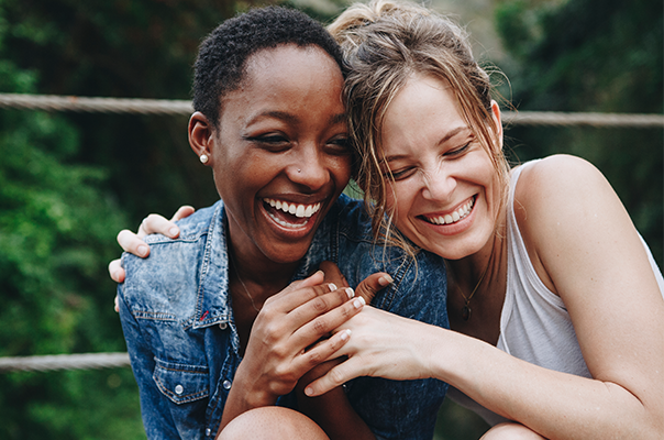 Zwei Frauen lachen fröhlich zusammen, während sie eng nebeneinander sitzen. Sie tragen lässige Kleidung und scheinen einen glücklichen Moment zu genießen. Im Hintergrund sind unscharfe grüne Bäume zu sehen, was eine natürliche Umgebung andeutet.
