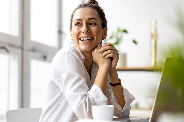 Eine lächelnde Frau in einem hellen Hemd sitzt an einem Tisch vor einem Laptop. Sie hat ihre Hände unter dem Kinn verschränkt und sieht freundlich aus. Eine Tasse steht vor ihr, und im Hintergrund sind Pflanzen und Regale zu sehen.
